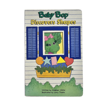 A children's book titled 'Baby Bop Discovers Shapes' with an illustration of a green dinosaur character looking out of a window with blue shutters, surrounded by cut-out shapes like stars and hearts.